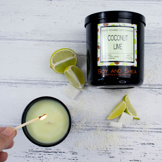 A flat lay photo showing a large black jar lying on its side on a white wooden surface.  To the bottom of the jar is a smaller candle standing upright and filled with light green wax.  A hand reaches in with a lit match.  Between the candles are wedges of lime and pieces of coconut flesh.