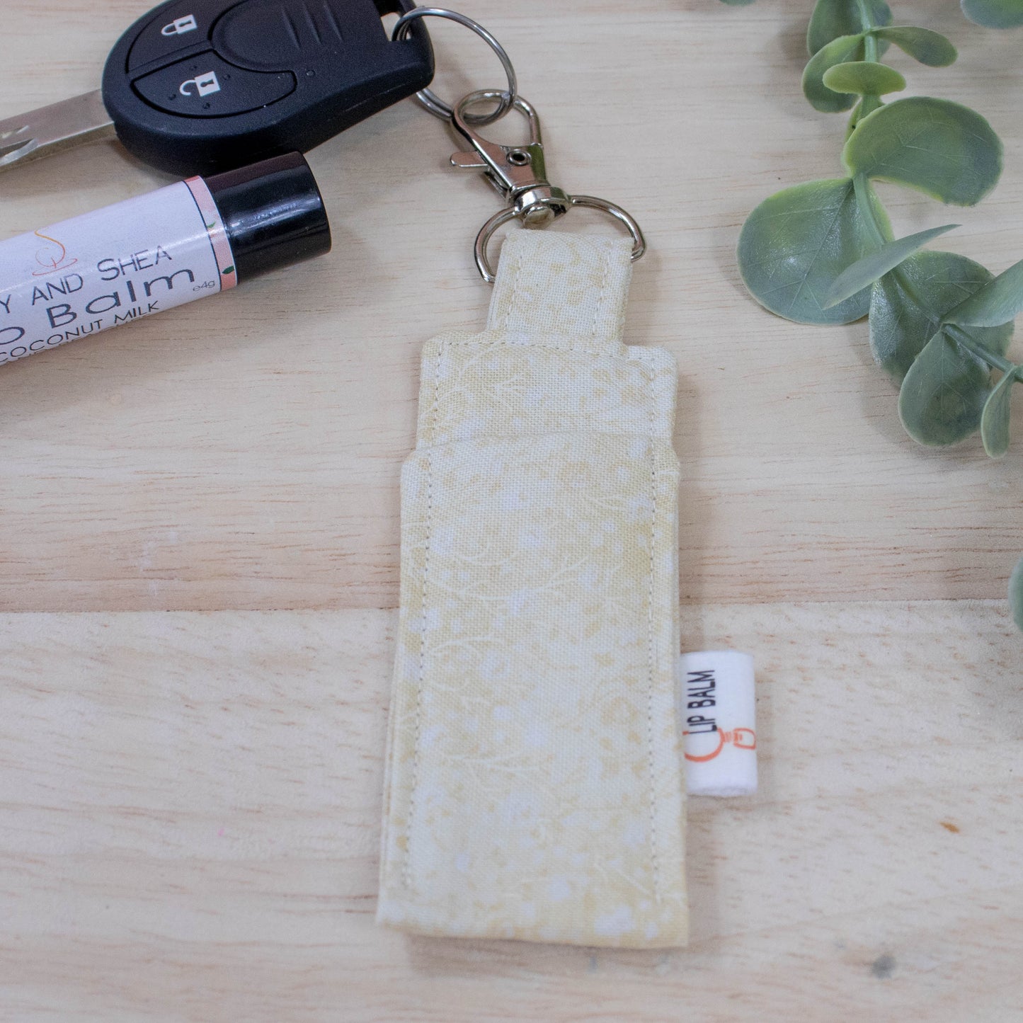 A clip balm holder lies on a wooden surface attached to a car key with a "lobster claw" clip.  To the side is a tube of lip balm and some greenery.  The Clip Balm is beige with a lighter pattern of flowres and leave like stems.