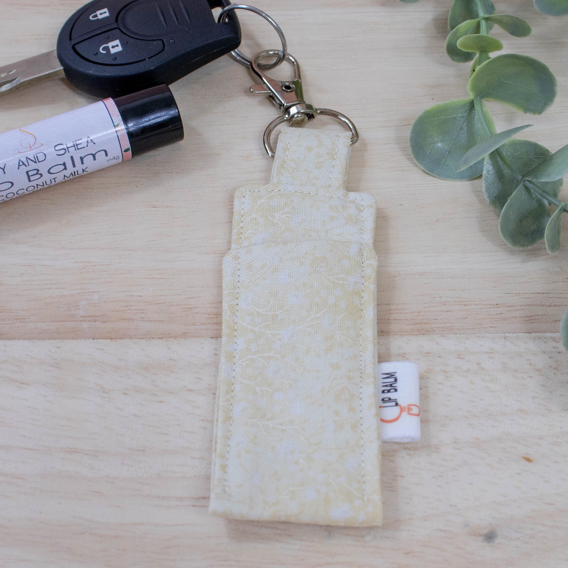 A clip balm holder lies on a wooden surface attached to a car key with a "lobster claw" clip.  To the side is a tube of lip balm and some greenery.  The Clip Balm is beige with a lighter pattern of flowres and leave like stems.