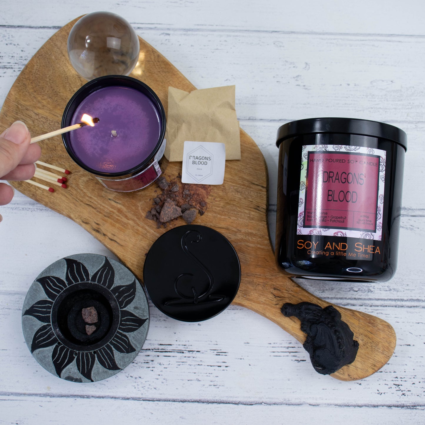 A flat lay photo showing a large black jar lying on its side on a white wooden surface next to a wooden tray where a smaller candle stands upright and filled with dark purple wax.  A hand reaches in with a lit match.  Surround the candle is some resin and a small bow along with a glass sphere.