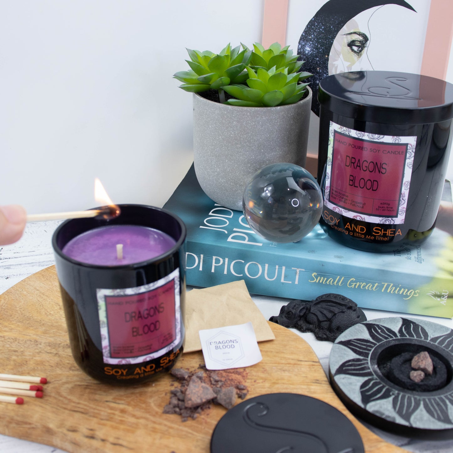 A candle in a glass black jar stands on a wooden surface next to a small pile of resin near a paper bag labelled dragons blood.  The metal lid from the candle lays near by with the logomark debossed into its surface. A lit match hovers over the candle whichhas a purple wax.  Behind lays a book which has a large candle sitting on top along with a potted plant and glass sphere.  A black dragon ornament and resin burn also appear.
