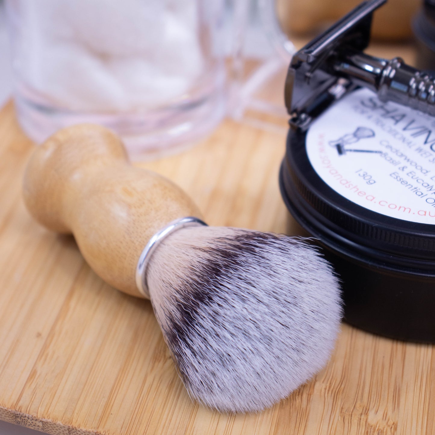 A close up image of a shaving brush laying flat on a wooden surface with its bristles pointing towards the camera next to a tin of shaving soap with a razor resting on top.  The handle of the brush is made of wood with silver neck collar and bristles in shades of cream, black and grey.