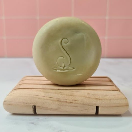 A round pale green soap stamped with logo sitting on a wooden soap tray