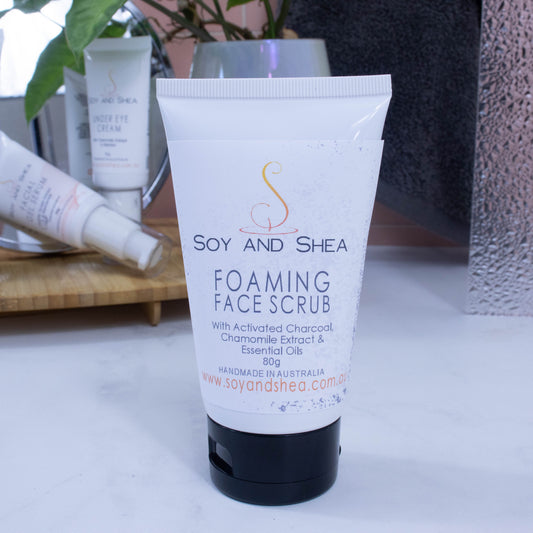 A white tube with black flip-top lids stands on a white surface against the background of a bathroom counter top displaying other facial products and greenery.  The tube has a white label with black flecks and is labelled FOAMING FACE SCRUB