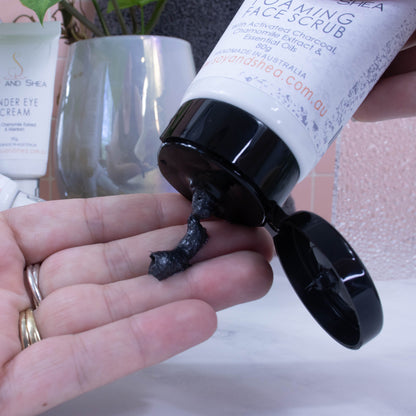 A hand is held out as a black soap is squeezed out of a white tube with black flip top lid with a label reading FOAMING FACE SCRUB.  Behind is a collection of items on a bathroom counter inclduing a potted plant and other bottles of face products.