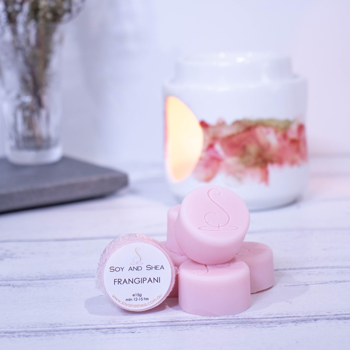 Four cylindrical shaped Melt Pucks in pale pink are stacked up on each other in pyramid form with the soy and shea logomark stamped on the tops.  To the left is a fourth melt puck that rests on its side with a label identifying it as Frangipani.  These sit on a wooden surface with a  lit fragrance warmer in the backgroung along with a vase of flowers.