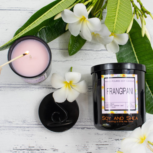 A flat lay photo showing a large black jar lying on its side on a white wooden surface.  To the left of the jar is a smaller candle standing upright and filled with light pink wax with a lit match hovering over the wick.  The candles are surrounded by bright green leaves and white frangipani flowers with yellow centers.  The black metal lid of the smaller candle lies between the two candles showing a debossed image of the logo mark.