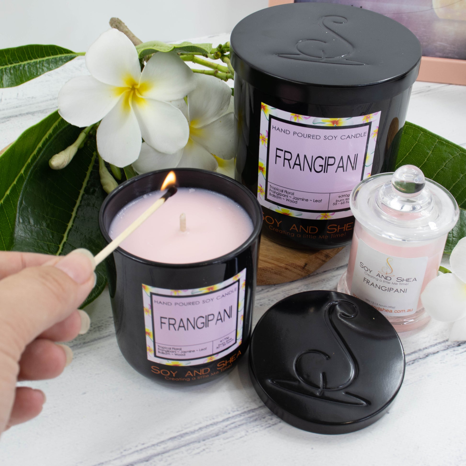 Two black glass candles are displayed on a white wooden surface along with a smaller candle in clear glass with light pink wax.  The candles are surrounded by white frangipanis with yellow centres and bright green leaves.  The larger of the two candles sits to the back with a black metal lid while a smaller one sits to the front with the lid removed to reveal a light pink wax.  A hand reaches in to light the candle with a match.  The lid of the candle sits to the front showing the debossed logomark image