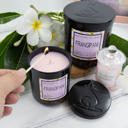 Two black glass candles are displayed on a white wooden surface along with a smaller candle in clear glass with light pink wax.  The candles are surrounded by white frangipanis with yellow centres and bright green leaves.  The larger of the two candles sits to the back with a black metal lid while a smaller one sits to the front with the lid removed to reveal a light pink wax.  A hand reaches in to light the candle with a match.  The lid of the candle sits to the front showing the debossed logomark image