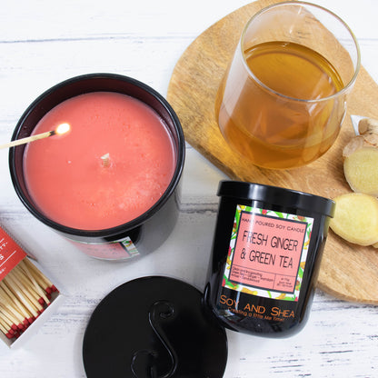 A flat lay photo showing a large black glass candle with red wax on a white wooden surface about to be lit with a match. Next to it, a small candle lies on its side revealng a decorative product label.  A tray holding a clear glass of tea and some slices of ginger appear above the candles.