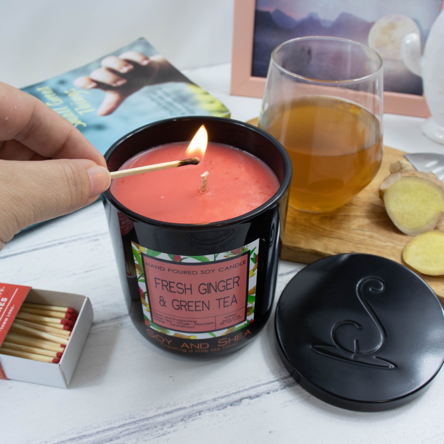 A hand reaches in with a lit match to light a candle in a black glass jar filled with red-orange wax.  Next to the jar lies a black metal lid with the logomark debossed in the center.  In the background is a clear glass of tea with slices of ginger beside it.  A book and watercolour painting are also in the background.