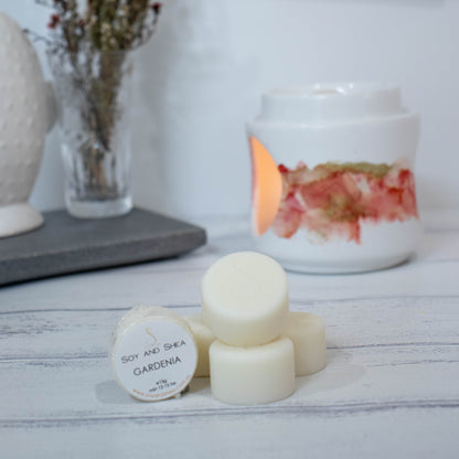 Four cylindrical shaped Melt Pucks in cream are stacked up on each other in pyramid form with the soy and shea logomark stamped on the tops.  To the left is a fourth melt puck that rests on its side with a label identifying it as Gardenia.  These sit on a wooden surface with a  lit fragrance warmer in the backgroung along with a vase of flowers.