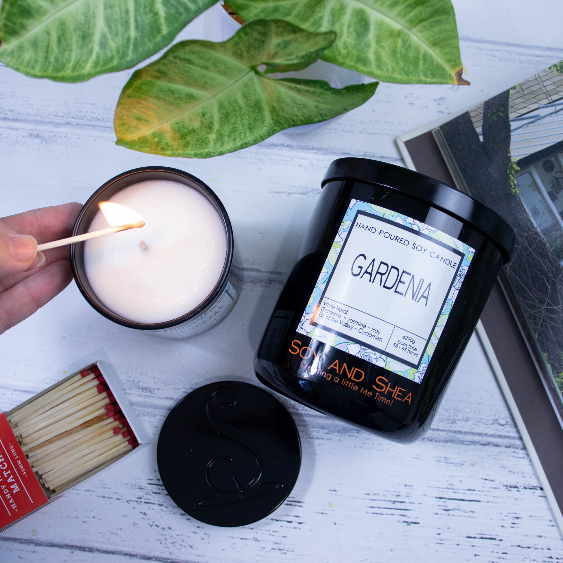 A flat lay photo showing a large black jar lying on its side on a white wooden surface next to a book and plant.  To the side of the jar is a smaller candle standing upright and filled with white wax.  A hand reaches in with a lit match.