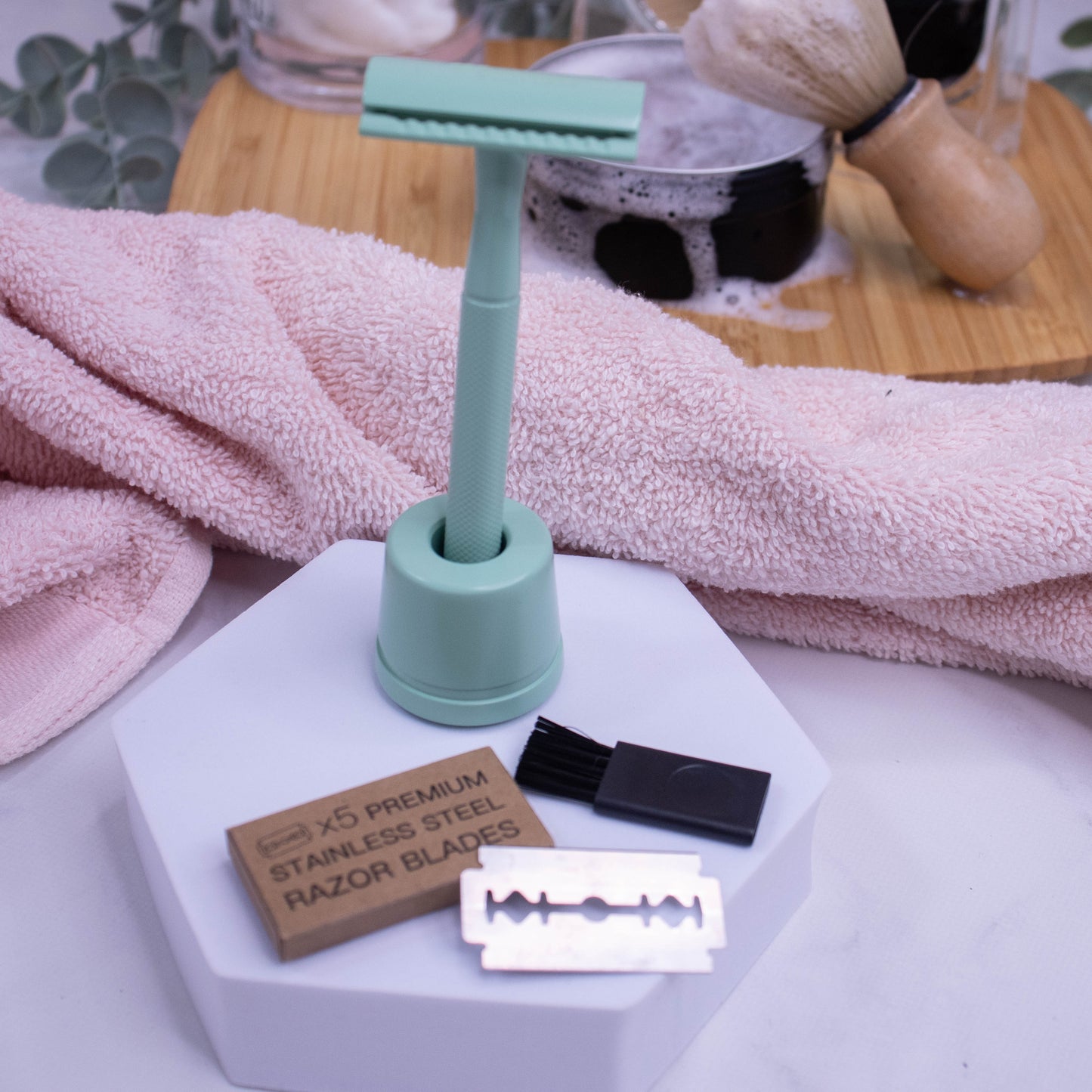 A pale green safety razor stands upright in its matching holder with a pack of five stainless steel blades and small brush in front on a white foam stand.  Behind is a pink towel and a tin of Shaving soap and a brush resting on a wooden tray.
