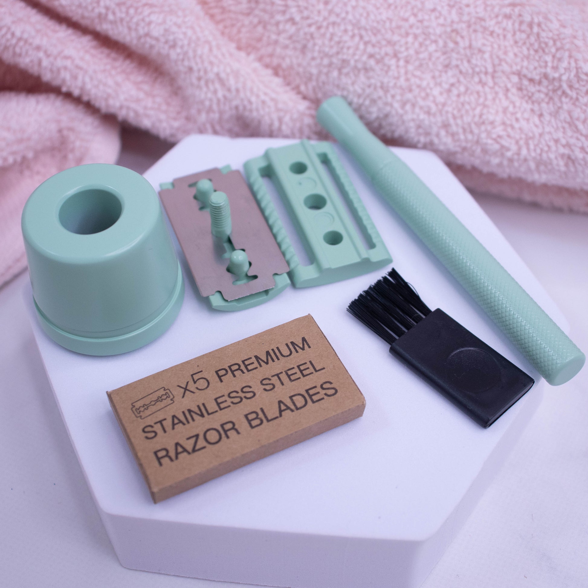 A pink towel lies by a white foam stand with a dismantled pale green safety razor.  It demonstates how the handle twists off the head of the razor which splits into two with a silver blade resting on one half of the head.  A pack of five razor blades in cardboard box and small black cleaning brush also rest on the white surface.