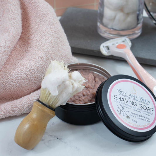 A black tin of shaving soap sits on a marble bench along with a pink razor and pink towel.  The lid has been removed and is resting on the side revealing a dusky pink soap inside.  A shaving brush with a dense foam rests against the other side of the tin.  The lid features a white round label with product detail and pink border.