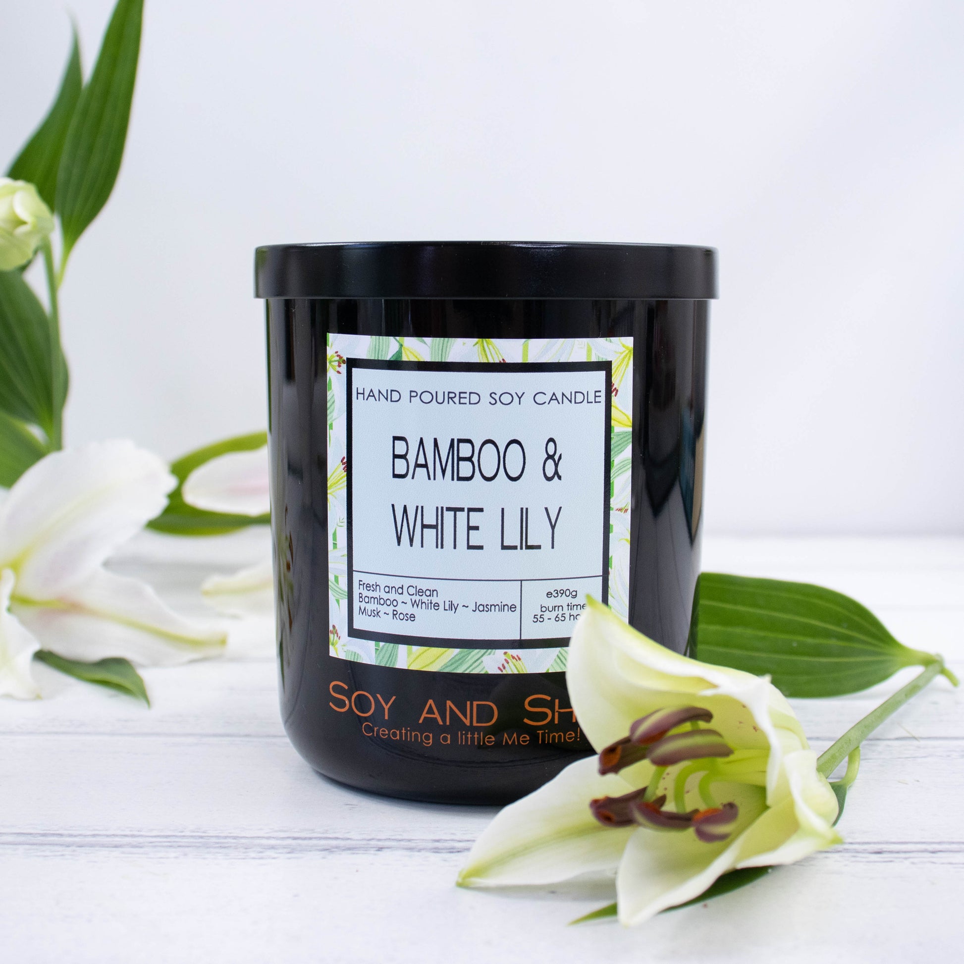 A black glass jar with black metal lid sits on the wooden side.  A decorative label is displayed on the front with information about the candle.  Surrounding the candle are stems of white lilies.