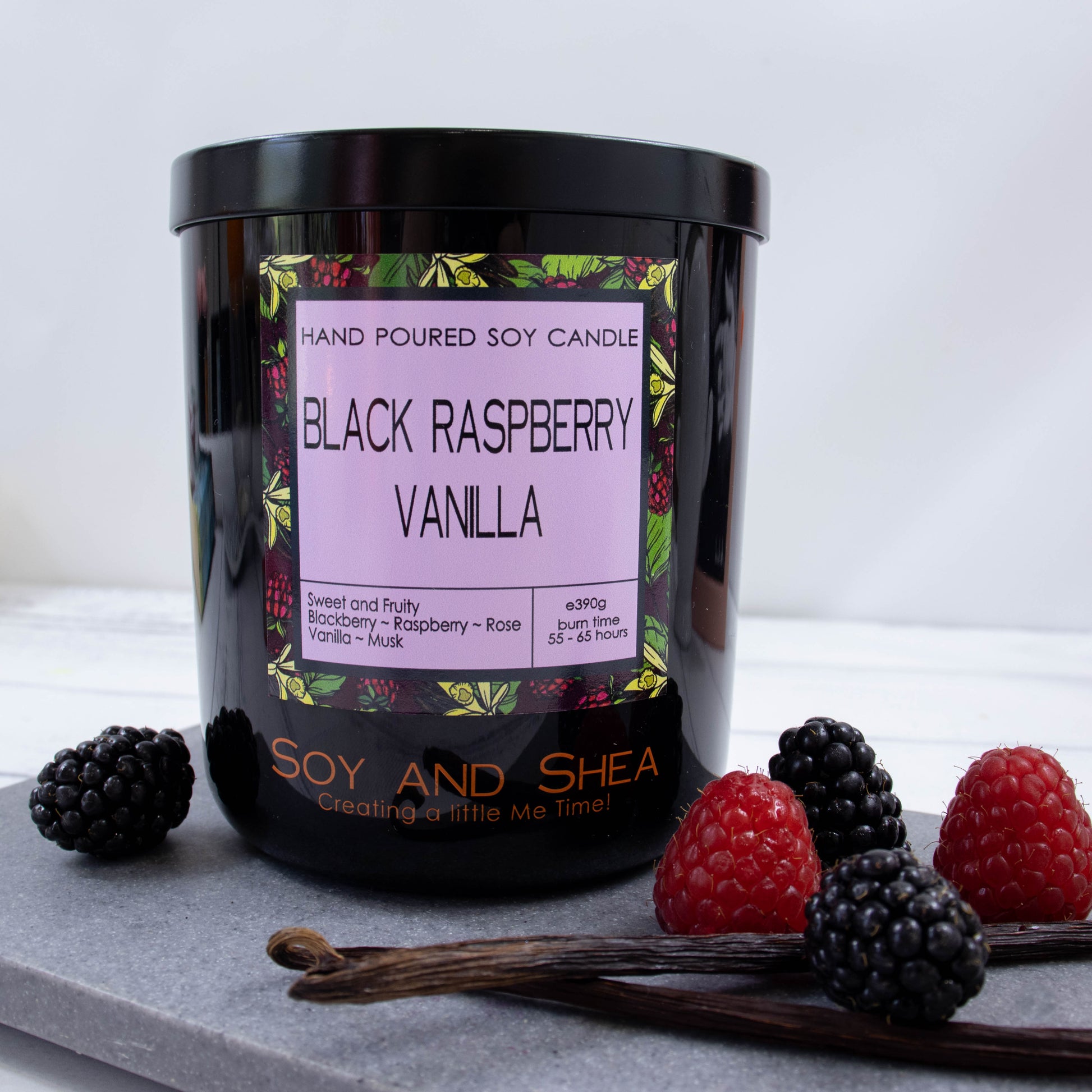 A black glass jar with black metal lid stands on the grey stone surface.  A decorative label is displayed on the front with information about the candle.   The candle is surrounded by a mix of black and red berries and two pods of vanilla.