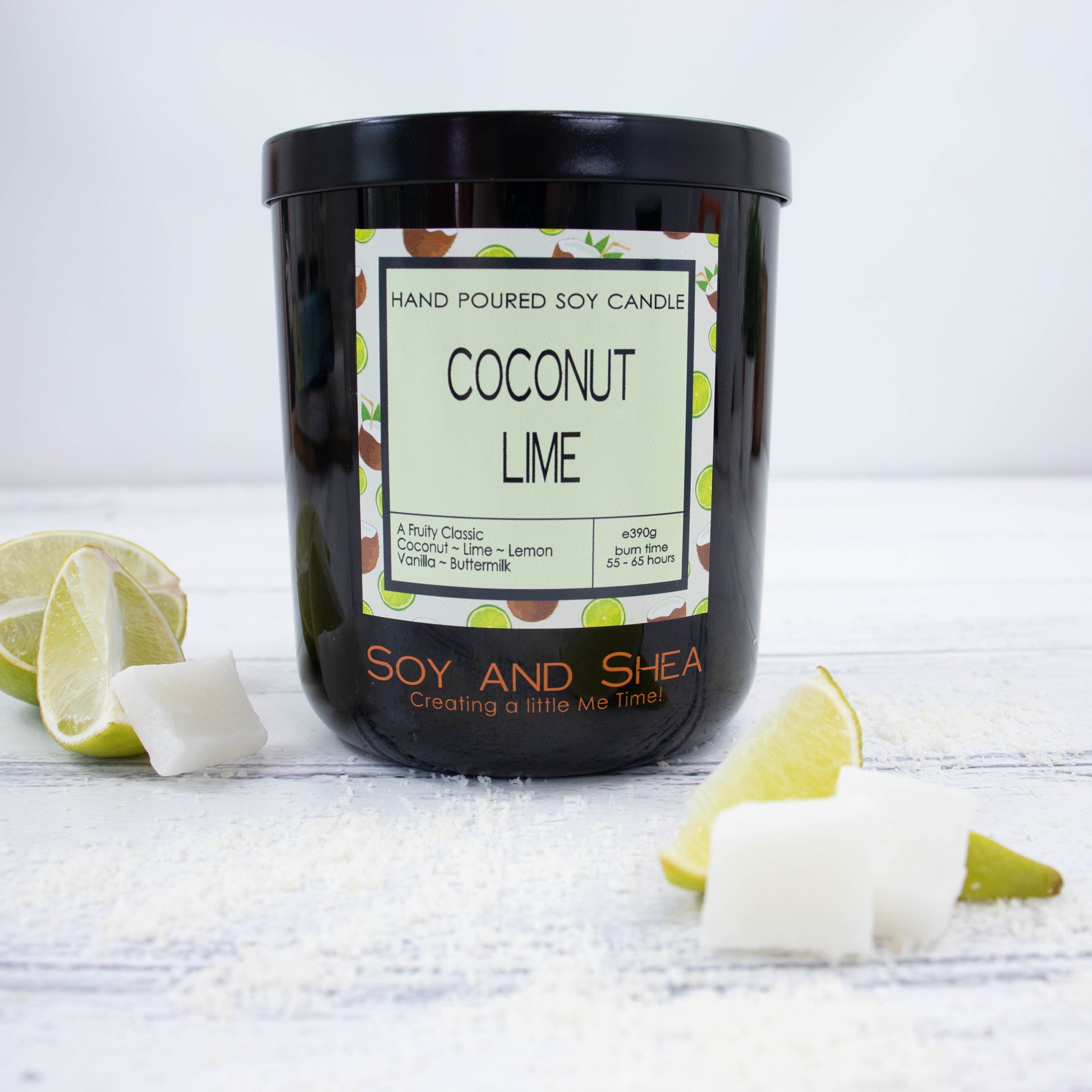 A black glass jar with black metal lid stands on the wooden side.  A decorative label is displayed on the front with information about the candle.   Surroundleing the candle are wedges of lime and piece of coconut flesh.