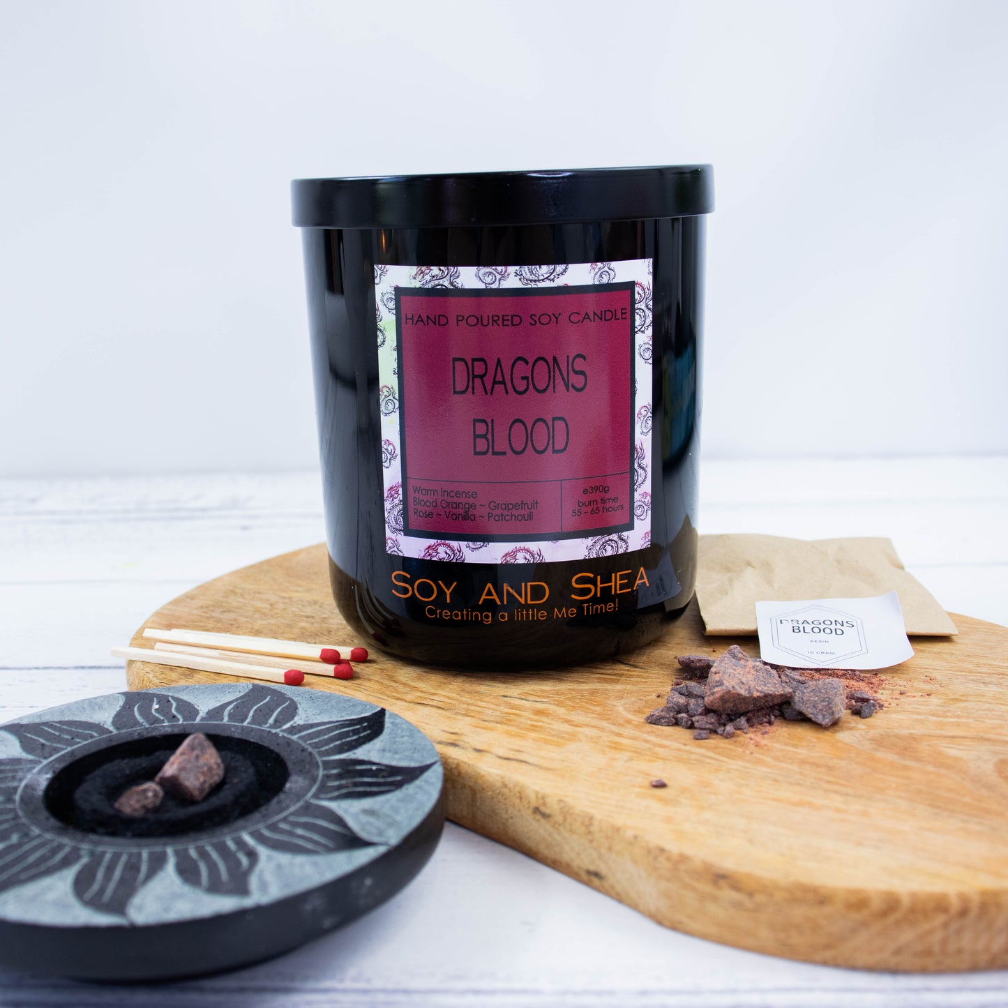 A black glass jar with black metal lid sits on the wooden side.  A decorative label is displayed on the front with information about the candle.  Surrounding the candle are pieces of resin with a paper bag labelled DRAGONS BLOOD along with a resin burner and matches.