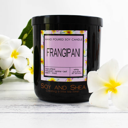 A black glass jar with black metal lid stands on the white wooden surface.  A decorative label is displayed on the front with information about the candle.   The candle is surrounded white franganpis with yelllow centres.