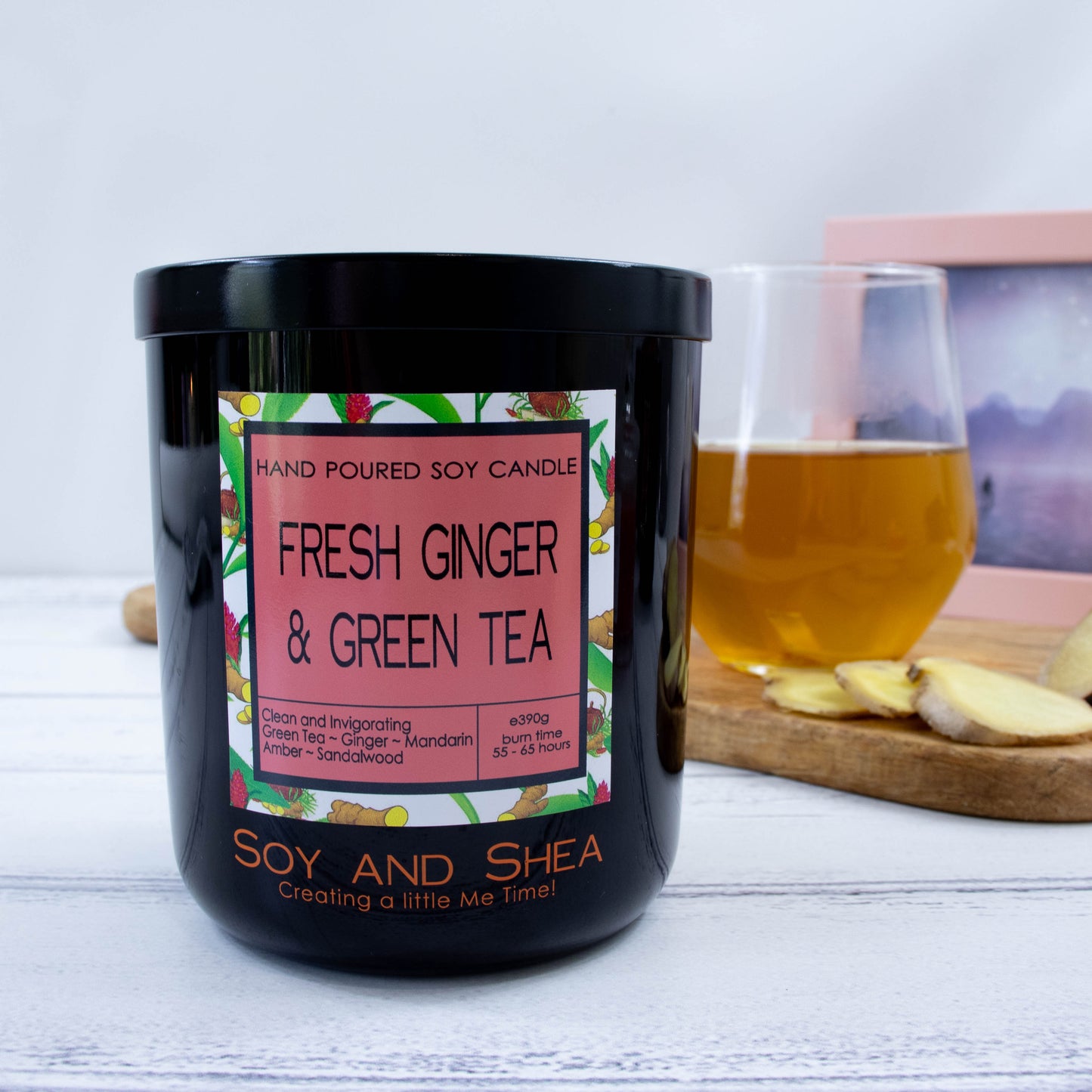 A black glass jar with black metal lid stands on the wooden side.  A decorative label is displayed on the front with information about the candle.   Behind the candle on a wooden tray is a a clear glass of tea along with some slices of ginger.