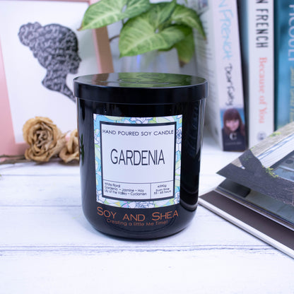 A large black glass jar with black metal lid sits on the wooden side.  A decorative label is displayed on the front with information about the candle.  In the background is a picture and some books.