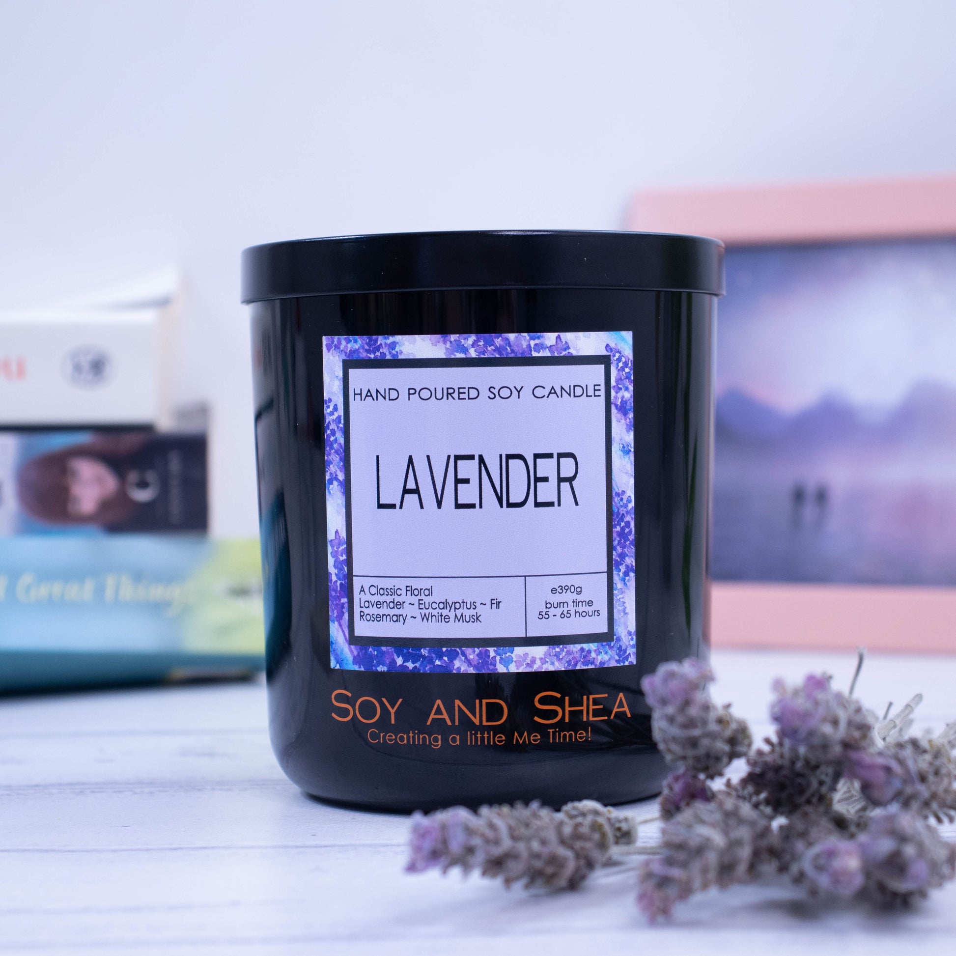 A large black glass jar with black metal lid sits on the wooden side surrounded by dry lavender flowers.  A decorative label is displayed on the front with information about the candle.  In the background is a picture and some books.