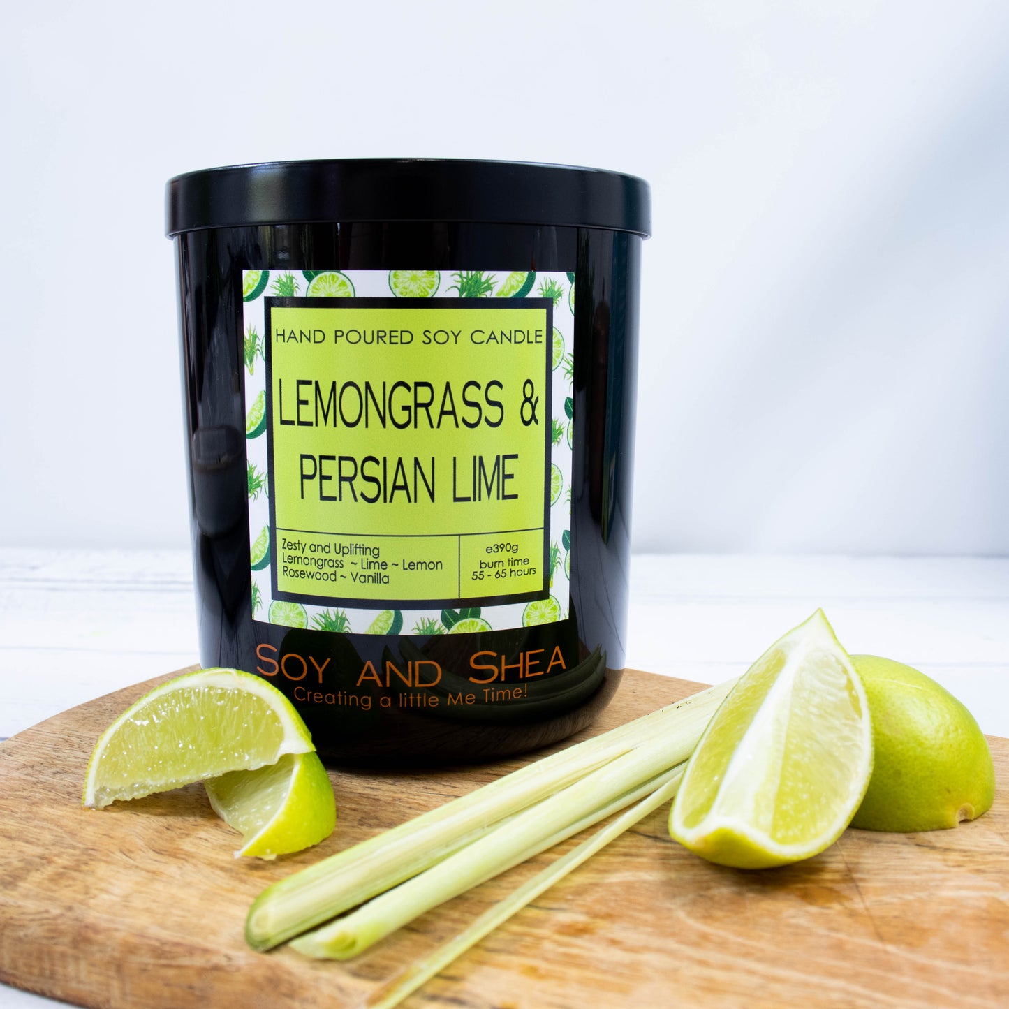 A black glass jar with black metal lid stands on the wooden side.  A decorative label is displayed on the front with information about the candle.   Beside the candle are some slices of lime and a stalk of lemongrass.
