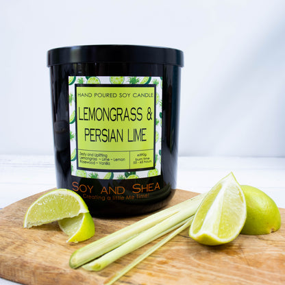 A black glass jar with black metal lid stands on the wooden side.  A decorative label is displayed on the front with information about the candle.   Beside the candle are some slices of lime and a stalk of lemongrass.