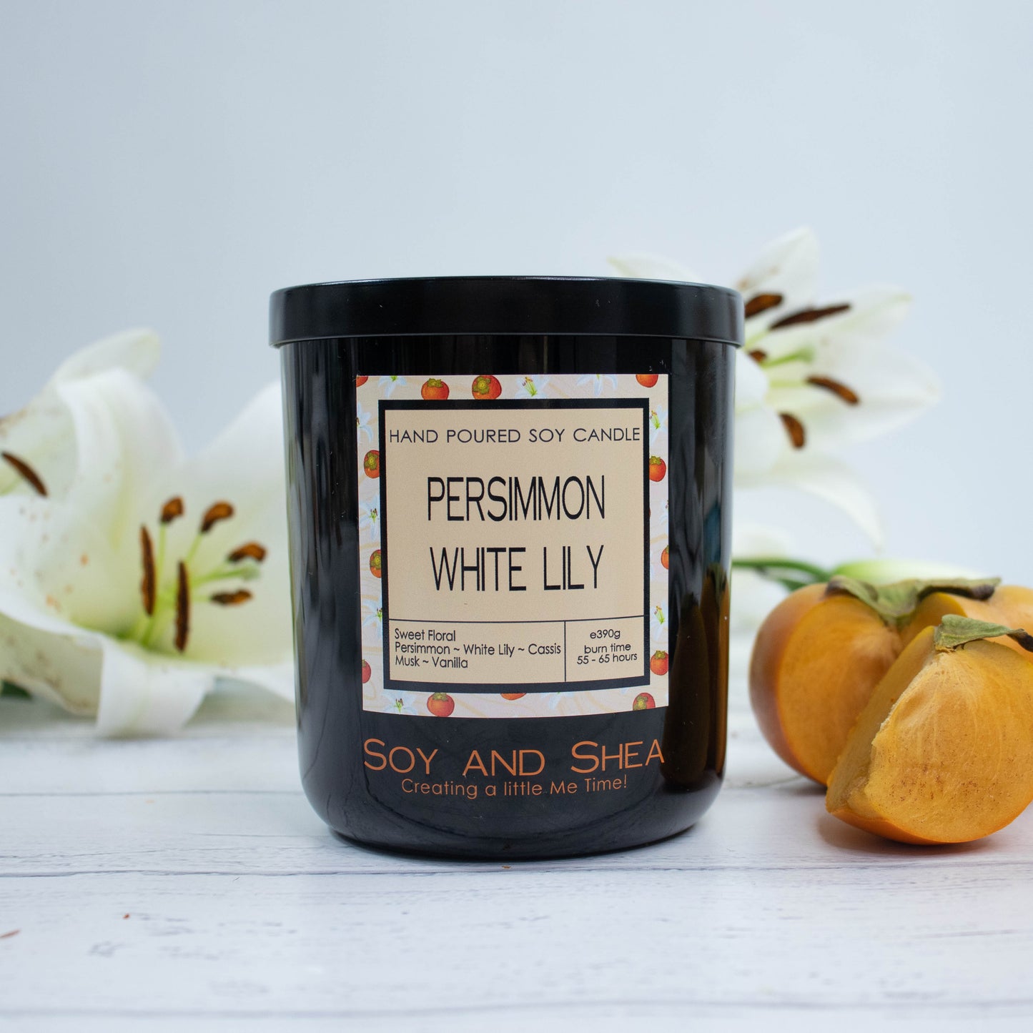 A black glass jar with black metal lid sits on the wooden side.  A decorative label is displayed on the front with information about the candle.  To the right of the candle is a ripe persimmon with a wedge cut out and behind are two white lily flowers.