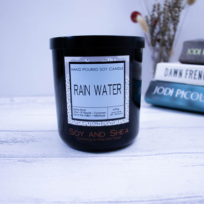 A black glass jar with black metal lid sits on the wooden side.  A decorative label is displayed on the front with information about the candle.  In the background is a stack of books besidea vase with dried flowers.