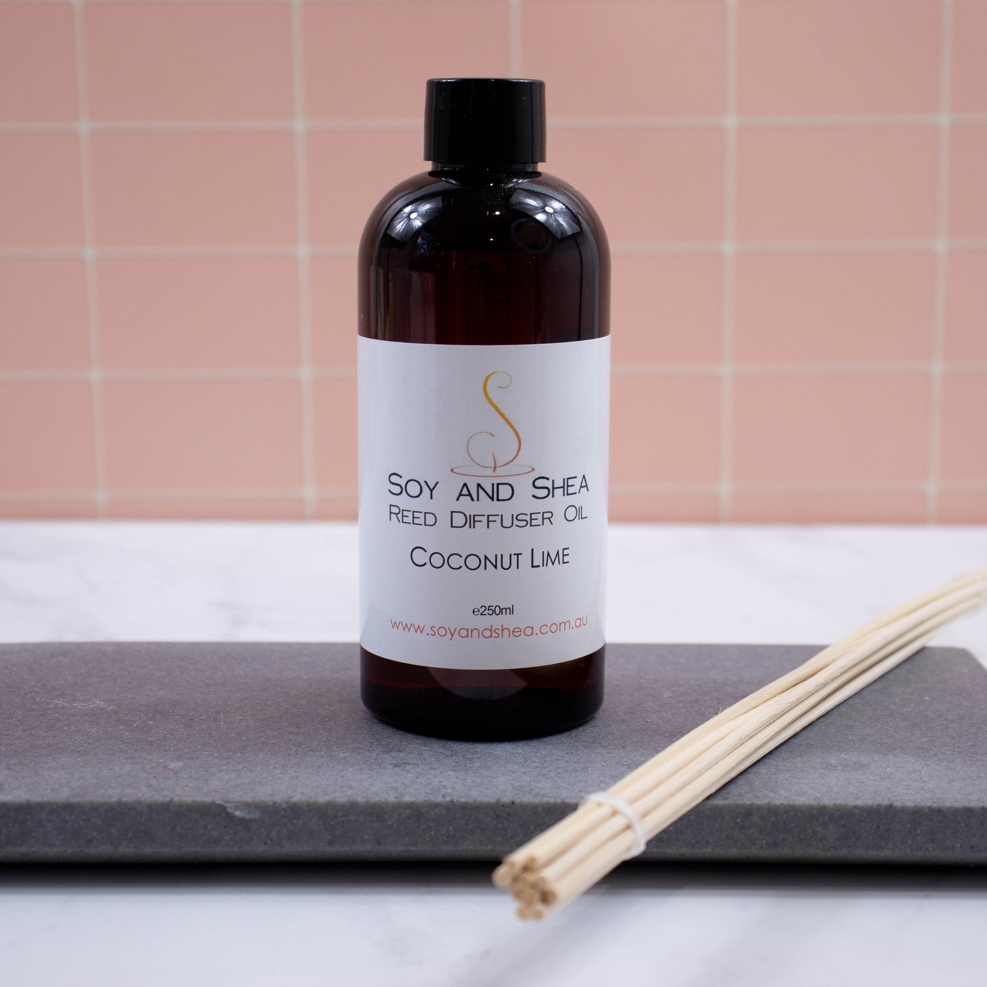 A brown bottle with black cap stands on a grey stone tray with a bundle of natural coloured reeds against a peach tile background.  It has a white label with text REED DIFFUSER OIL 250ml
