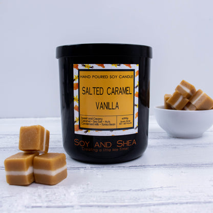 A black glass jar with black metal lid sits on the wooden side.  A decorative label is displayed on the front with information about the candle.  To the left of the candle is a three caramel lollies with a small pile of salt.  In the background is white bowl filled with caramels.