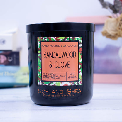 A large black glass jar with black metal lid sits on the wooden side.  A decorative label is displayed on the front with information about the candle.  In the background is a picture and some books.