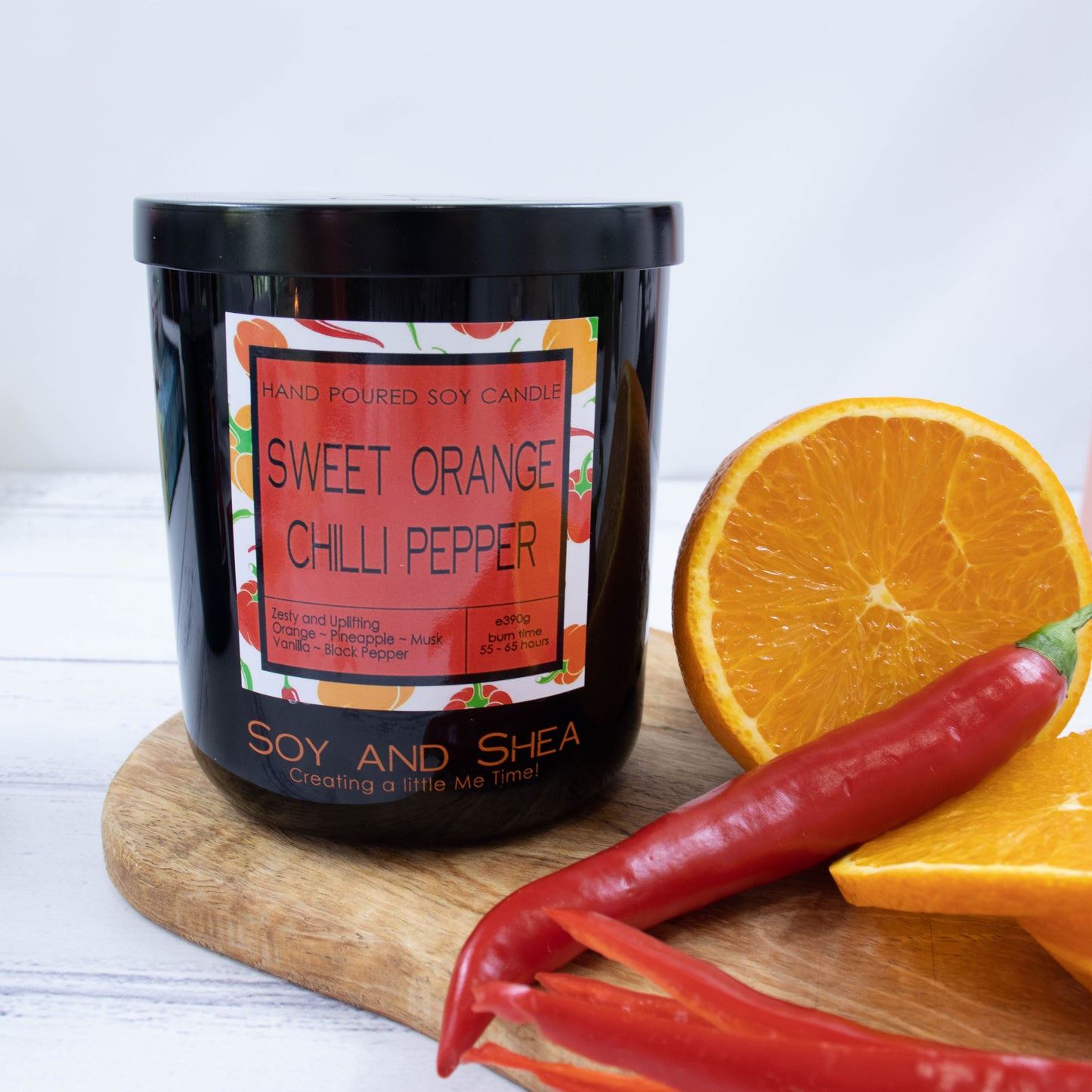 A black glass jar with black metal lid stands on the wooden side.  A decorative label is displayed on the front with information about the candle.   Beside the candle sits an orange that has been cut in half and some red chillies.