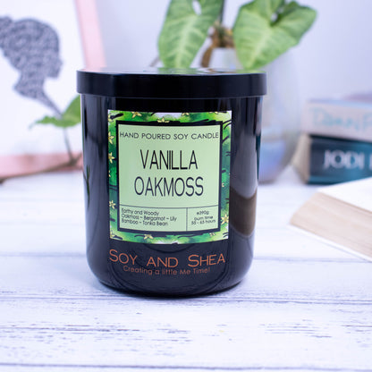 A large black glass jar with black metal lid sits on the wooden side.  A decorative label is displayed on the front with information about the candle.  In the background is a picture and some books.