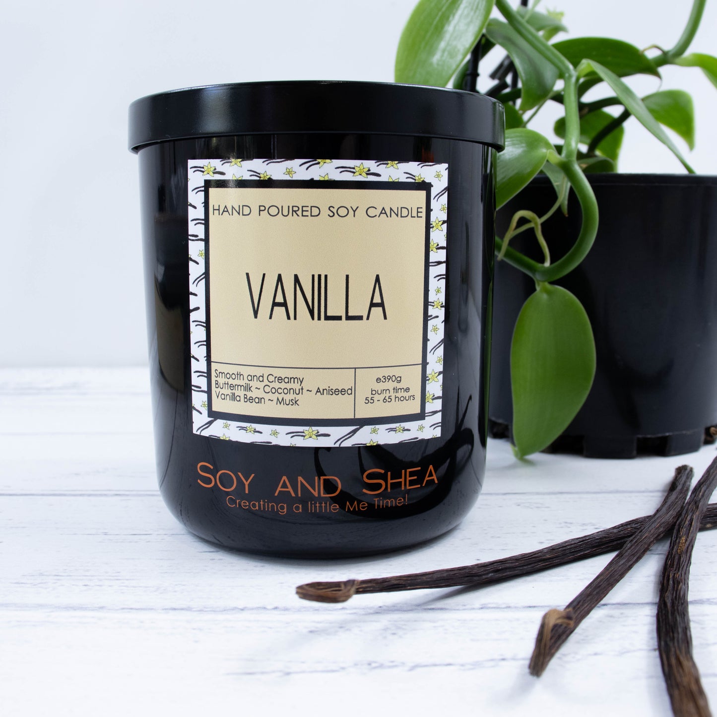 A black glass jar with black metal lid stands on the wooden side.  A decorative label is displayed on the front with information about the candle.   Beside the candle lies three dried vanilla  pods and a potted vanilla vine can be seen in the background.