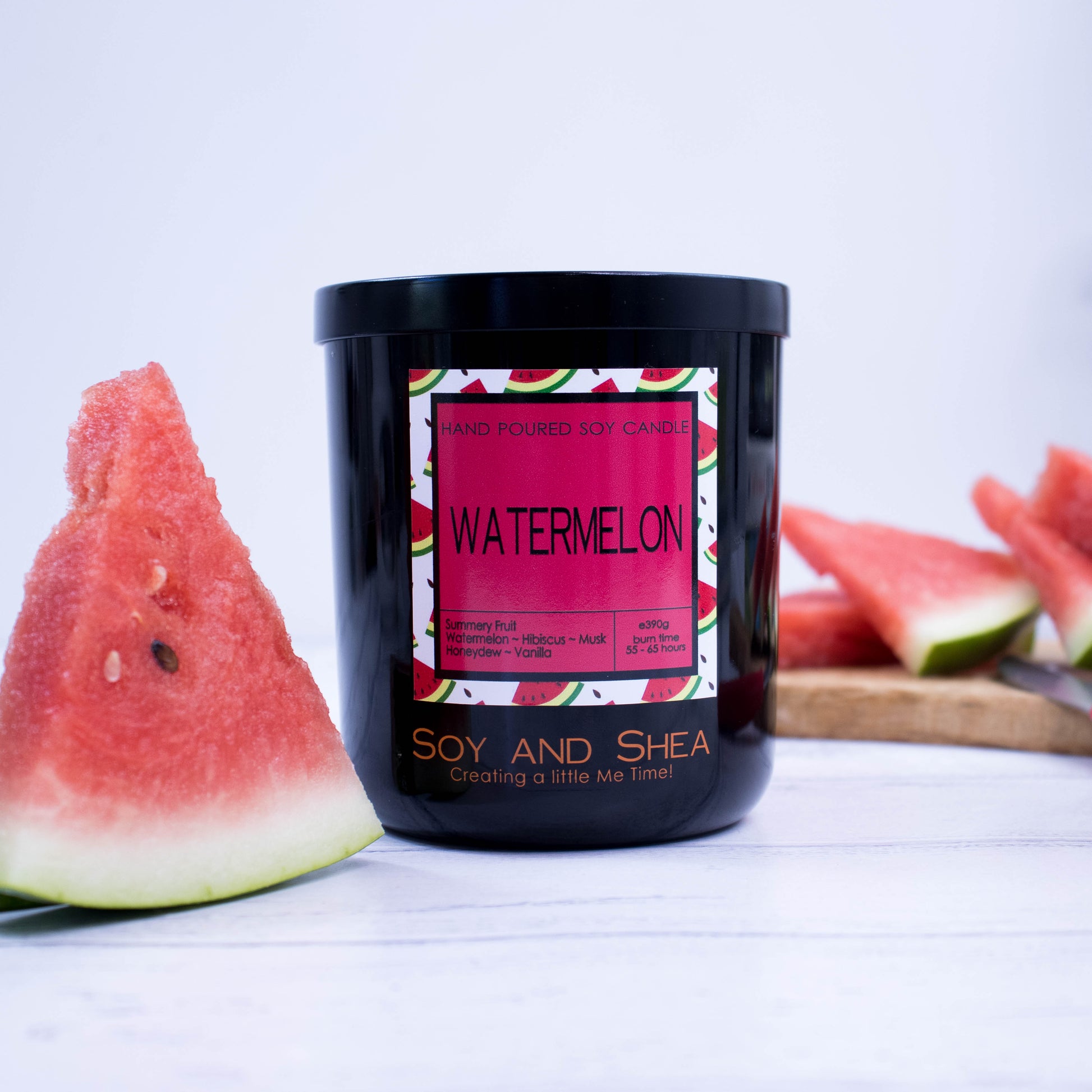 A black glass jar with black metal lid sits on the wooden side.  A decorative label is displayed on the front with information about the candle.  To the left of the candle is a piece of watermelon standing upright and in the blurred background is a wooden tray with more watermelon slices.