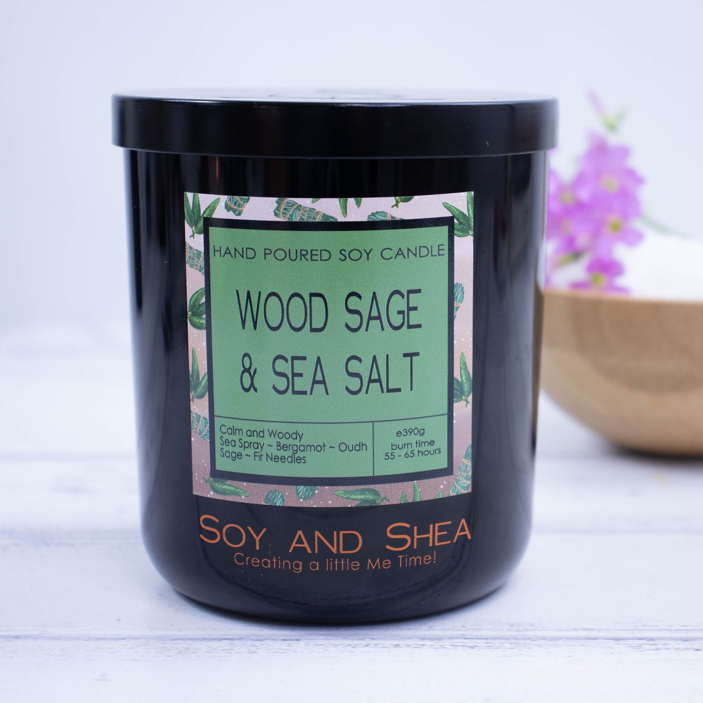 A Black jar with colourful product label and black metal lid stands on a wooden surgface with bright white background.  A wooden bowl with purple flowers sit behind the candle.