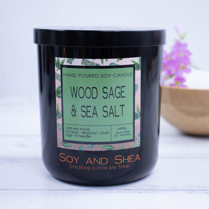 A Black jar with colourful product label and black metal lid stands on a wooden surgface with bright white background.  A wooden bowl with purple flowers sit behind the candle.