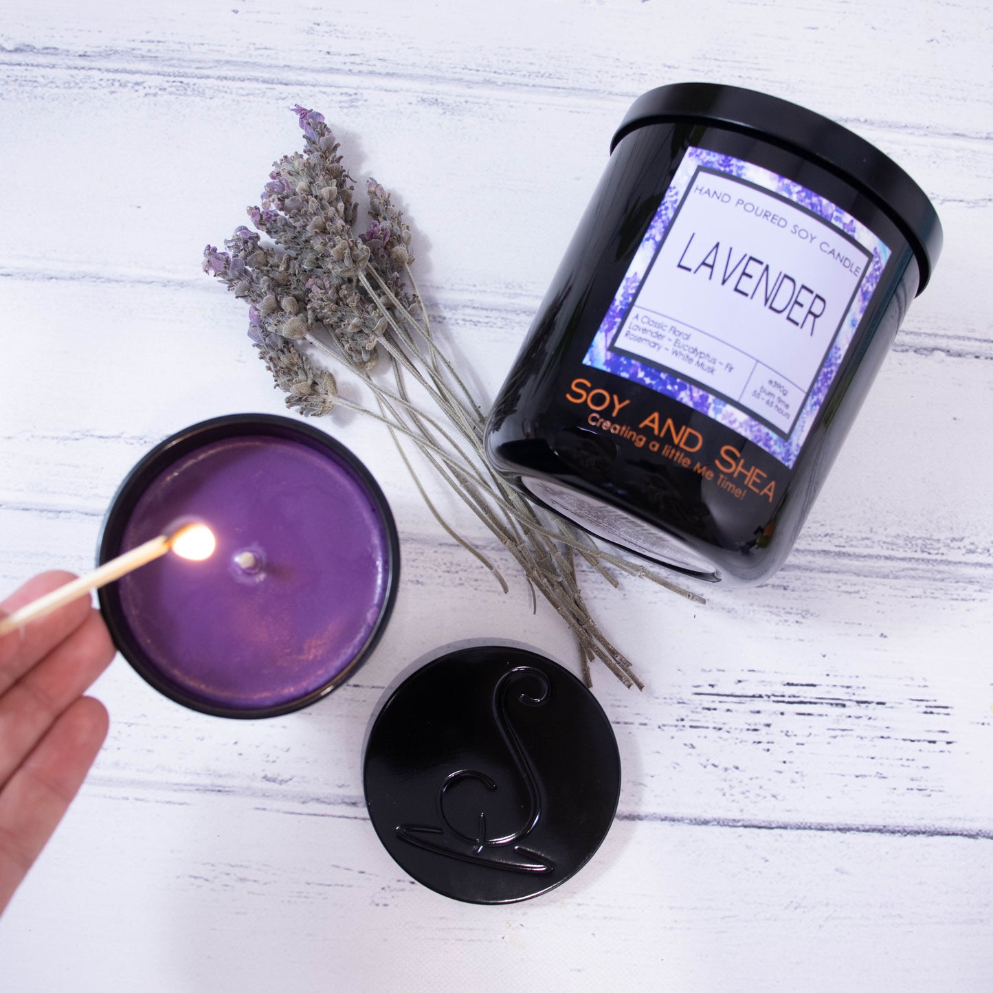 Two candles  in black jars are on a wooden bench.  The larger candle lies on it side with the label facing up.  A small candle sits up-right filled with purple wax.  To its side is a black metal lid and a hand is about to light the candle with a match.  Between the jars is a bunch of dried lavender