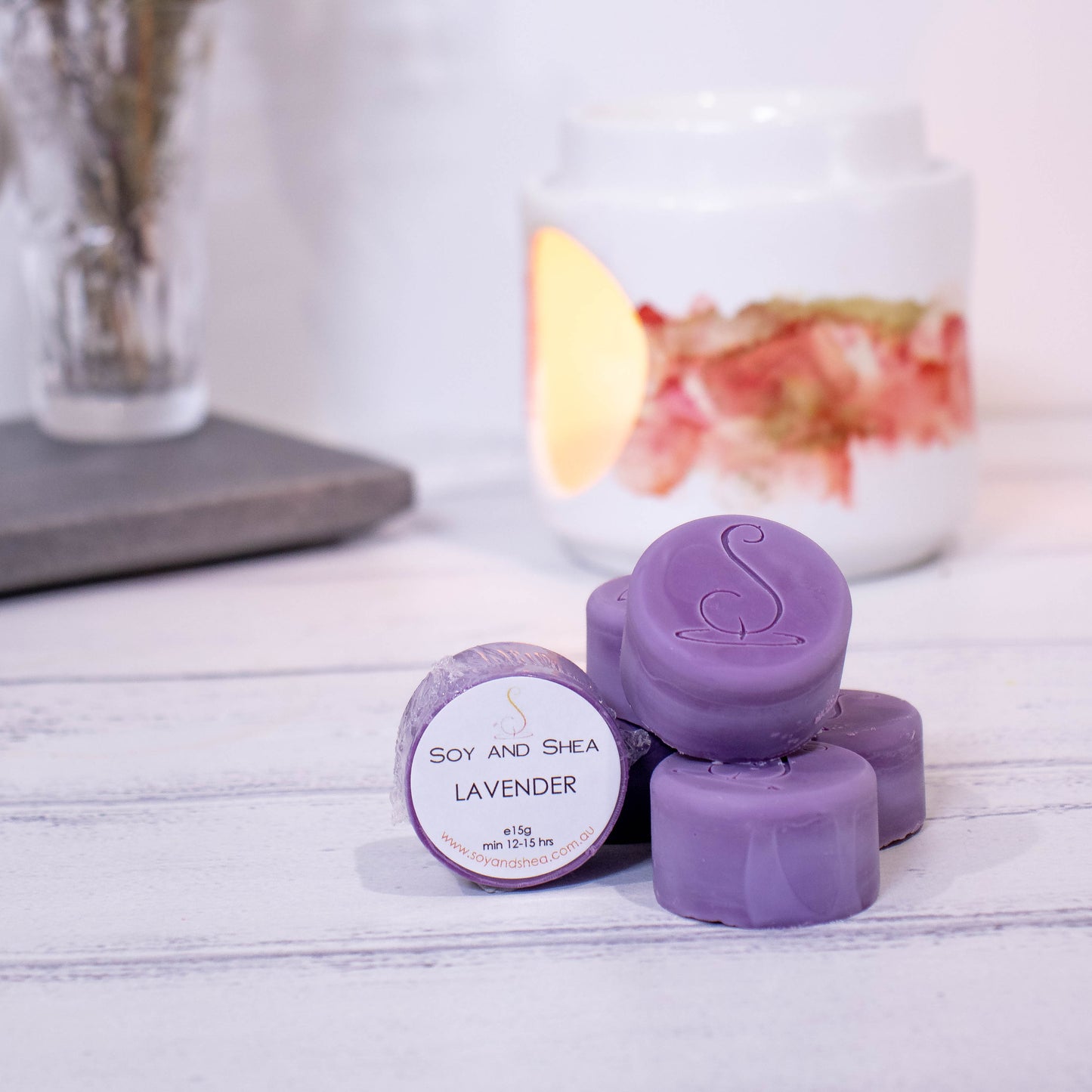 Four cylindrical shaped Melt Pucks in dark pruple are stacked up on each other in pyramid form with the soy and shea logomark stamped on the tops.  To the left is a fourth melt puck that rests on its side with a label identifying it as Lavender.  These sit on a wooden surface with a  lit fragrance warmer in the backgroung along with a vase of flowers.