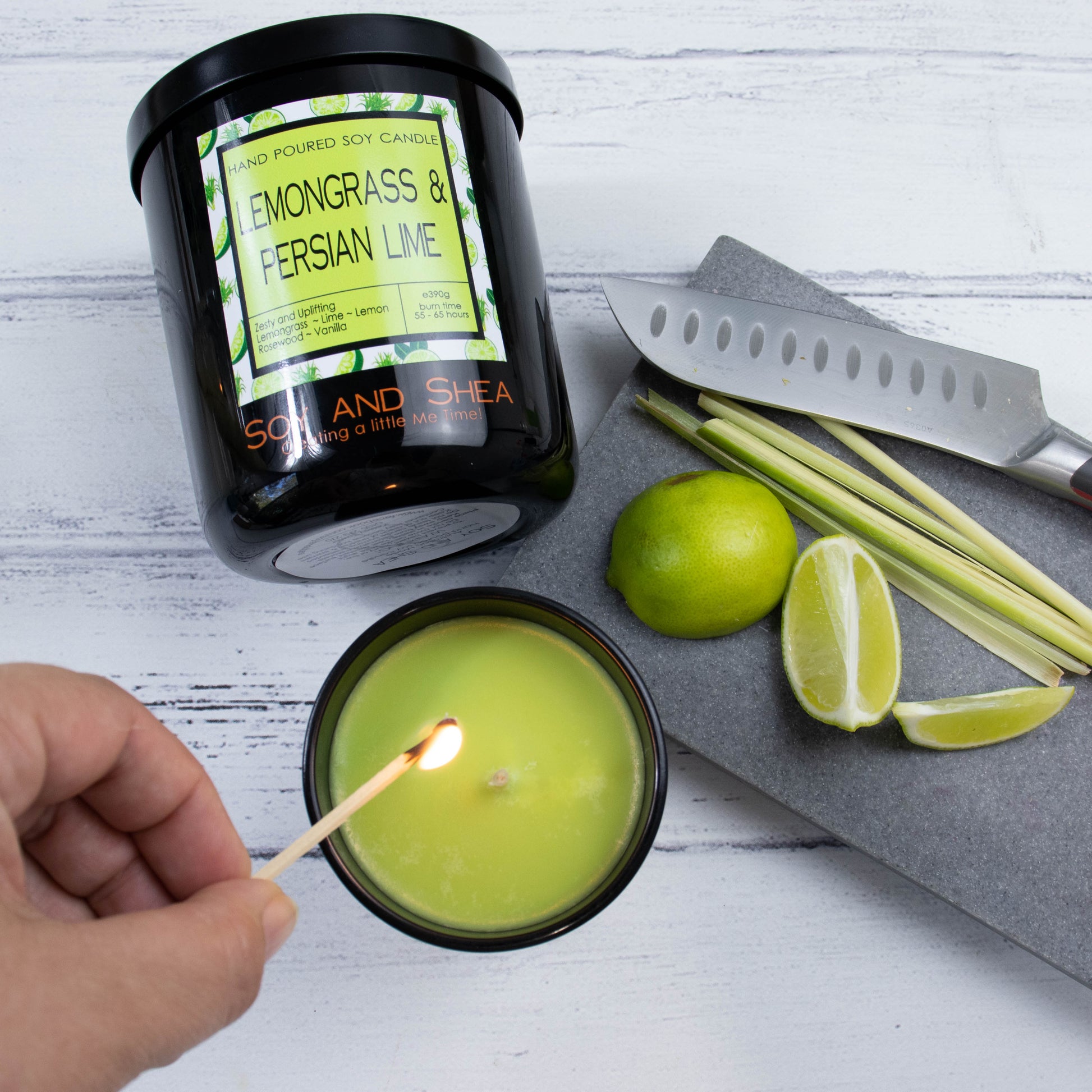 A flat lay photo showing a large black jar lying on its side on a white wooden surface.  To the bottom of the jar is a smaller candle standing upright and filled with bright green wax with a hand reaching in with a lit match.  To the right is a stone tray holding wedges of lime, stalks of lemongrass and a knife.