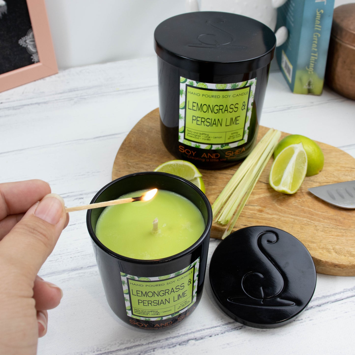 A hand reaches in to light a medium black glass candle with bright green wax.  Next to it on white wooden surface lies the black metal lid with the logomark debossed in the centre.  Behind is a wooden board with wedges of lime and a stem of lemongrass along with a larger version of the candle but with its lid on.  Books can be seen in the background.