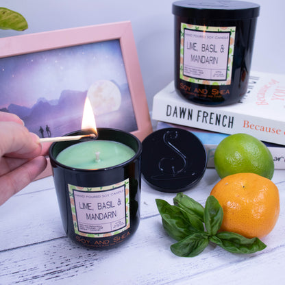 A small black candle filled with green wax and being lit with a match, stands on a wooden surface next to a mandarin, lime and bunch of basil.   The lid rests against a stack of books in the background which has a larger candle standing on top of them. A photoframe with a watercolour artwork appears in the background.