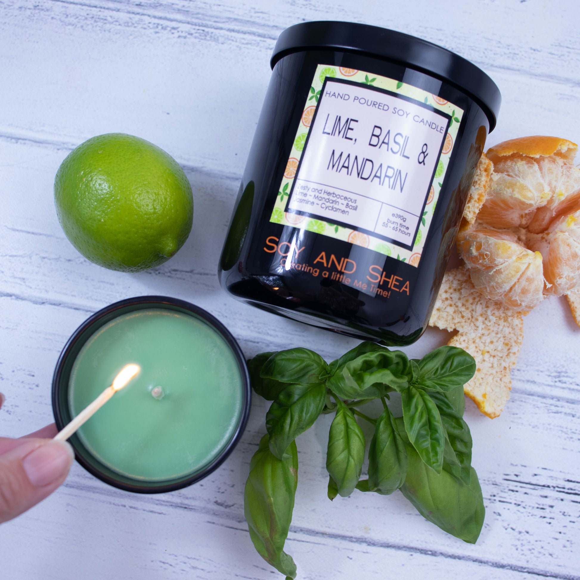 A flat lay photo showing a large black jar lying on its side on a white wooden surface surrounded by a whole lime, peeled mandarin and basil leaves.  To the bottom of the jar is a smaller candle standing upright and filled with green wax.  A hand reaches in with a lit match.
