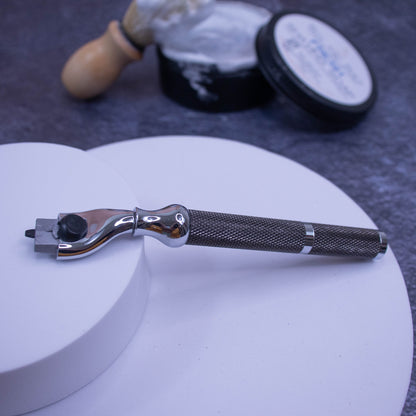 A dark silver Parker Cartridge Razor Handle (42R), compatible with MACH3 blades, rests on a white platform. In the blurred background are a shaving brush, shaving cream, and a dark container on a gray surface.