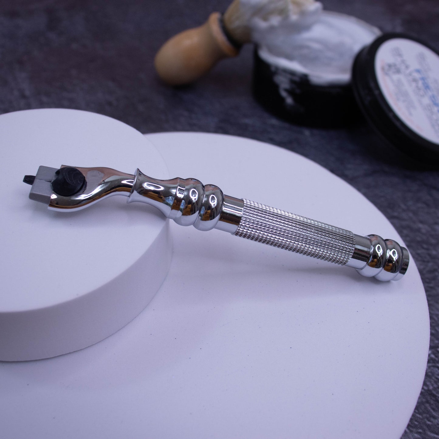 A shiny silver Parker Cartridge Razor Handle (43R), compatible with MACH3 blades, rests on a white platform. In the blurred background are a shaving brush, shaving cream, and a dark container on a gray surface.