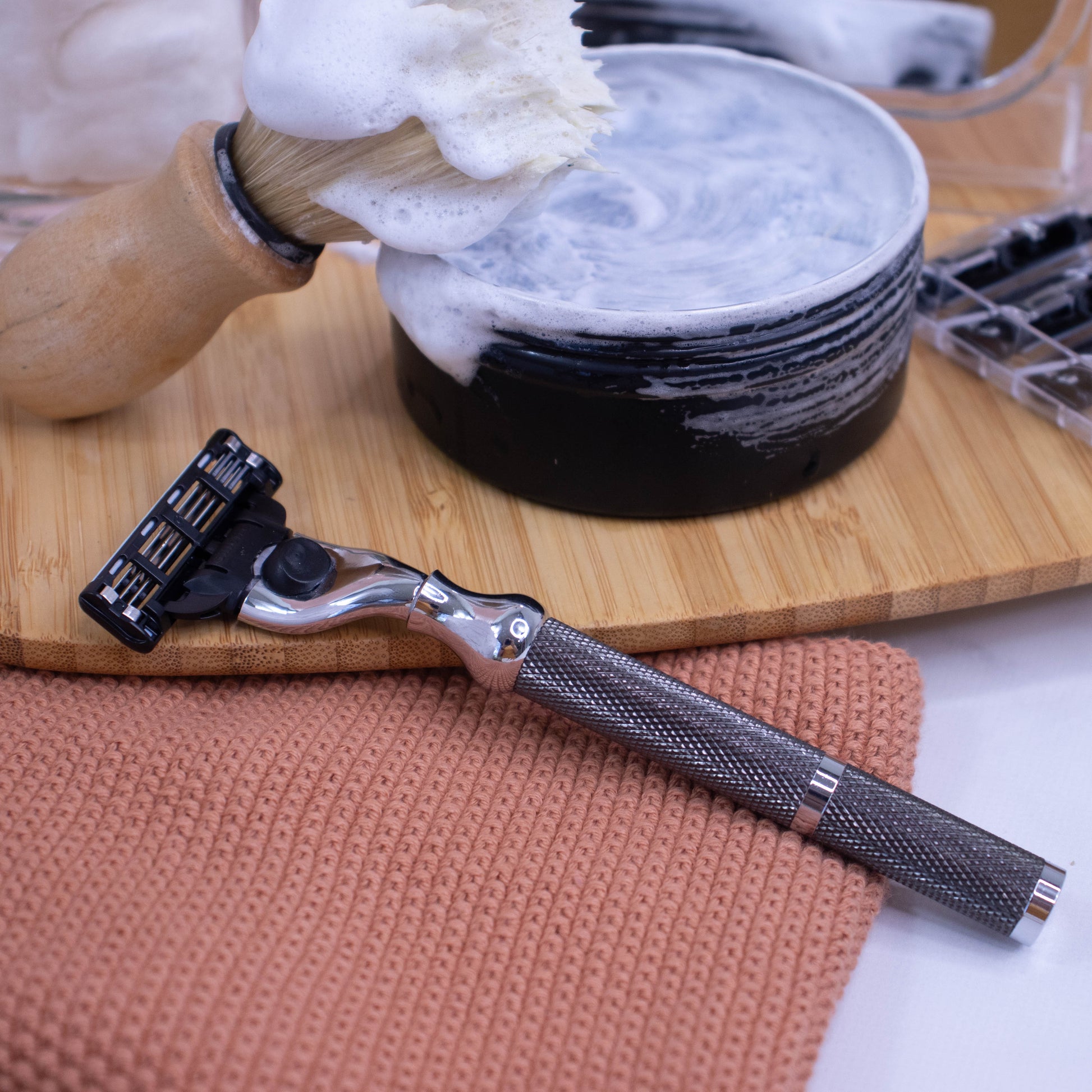 A dark silver-colored Parker cartridge razor handle with an all-metal textured grip displayed resting against a wooden tray with a close-up of the handle showing the chrome plating and a cartridge razor head.  Behind sits a tin of shaving soap with a shaving brush covered in the soap lather against a blurred bathroom background.